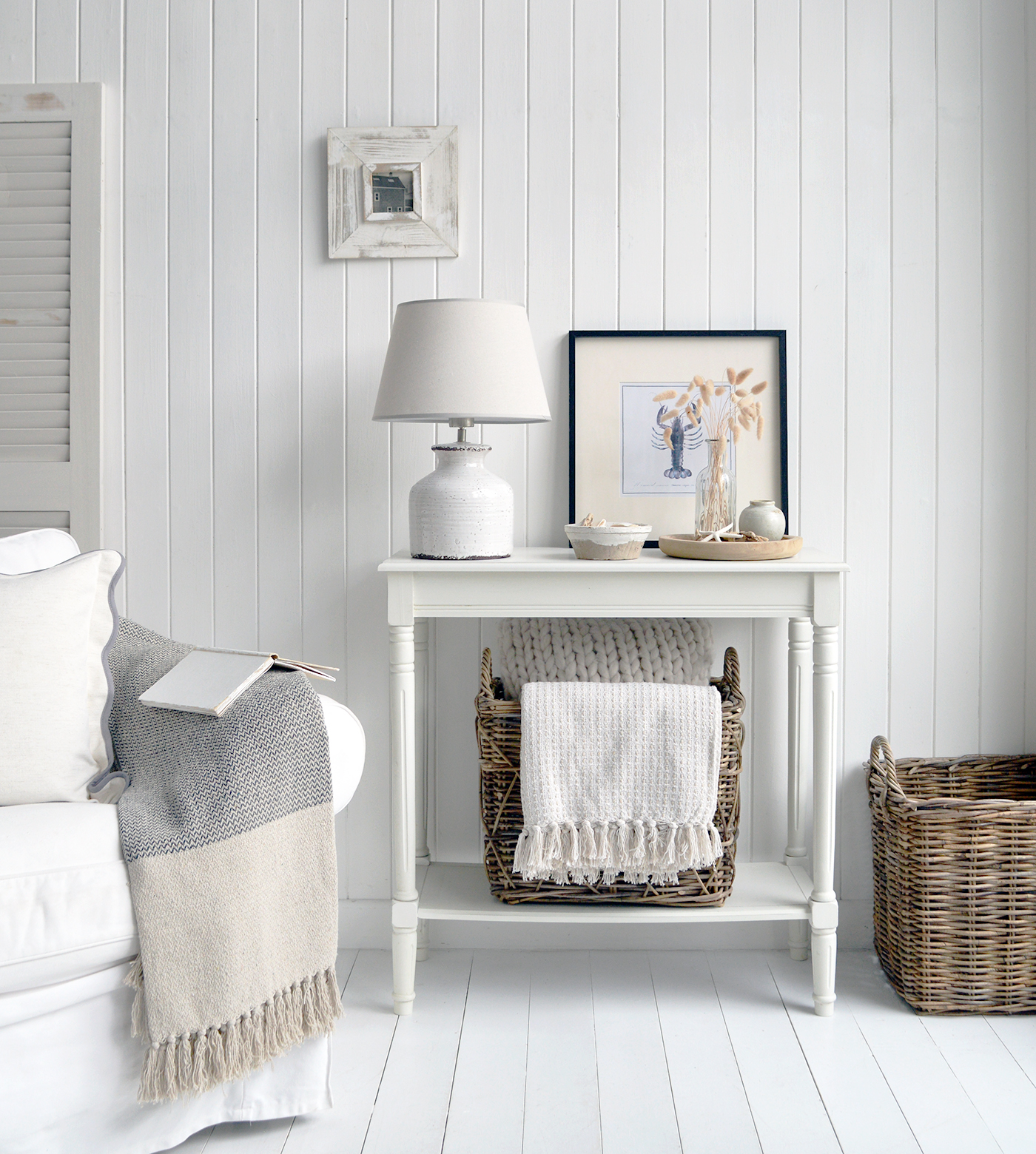 A white console table in classic Hamptons seaside style stands elegantly against a light, airy wall. Its clean lines and crisp finish evoke coastal sophistication. On the lower shelf, a woven basket overflows with soft throws and linen cushions in muted blues and sandy neutrals, adding texture and inviting comfort. Above, a framed coastal print—perhaps a sailboat or serene shoreline—anchors the vignette, completing the relaxed, refined look.